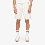 A close-up shot showcasing a model wearing off-white casual shorts with a drawstring waistband. The shorts are paired with white mid-calf socks featuring black stripes and white sneakers with beige soles, creating a clean and sporty aesthetic. The outfit emphasizes comfort and style with minimalist details