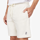 This image shows a close-up of the off-white shorts worn by the model. The shorts feature a drawstring waist, and the model has one hand in the pocket while the other rests on the side. On the bottom right corner of the shorts, there's a small embroidered logo of a dog wearing a helmet, adding a playful touch to the design