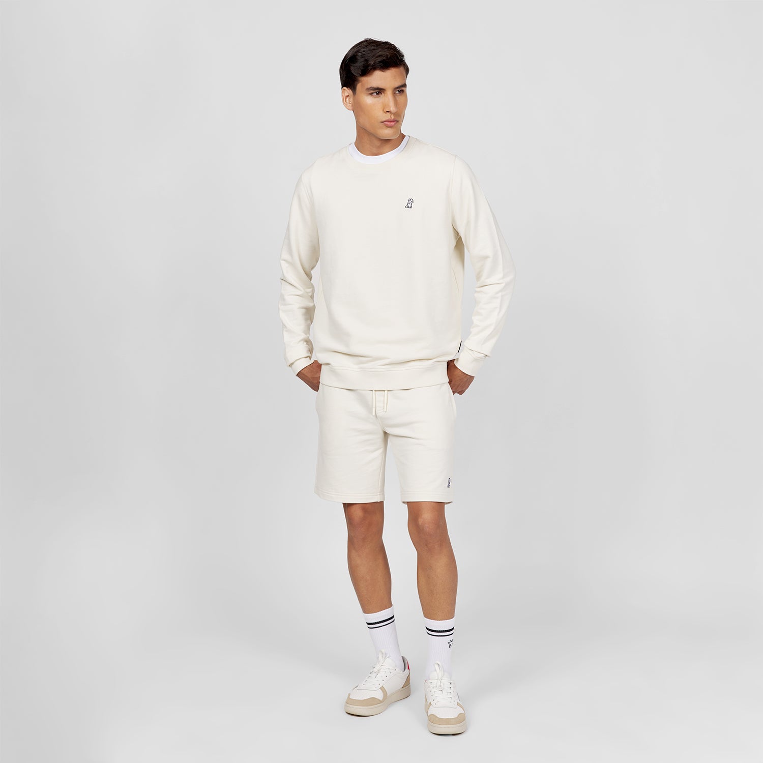 This image shows the model wearing a casual and comfortable off-white outfit consisting of a crewneck sweatshirt and matching shorts. The sweatshirt features a small embroidered logo on the chest, and the shorts have a similar embroidered logo on the bottom, giving the outfit a clean and coordinated look. The model also wears sneakers and white socks with black stripes, completing the relaxed, sporty style