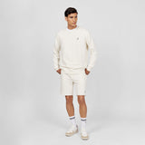 This image shows the model wearing a casual and comfortable off-white outfit consisting of a crewneck sweatshirt and matching shorts. The sweatshirt features a small embroidered logo on the chest, and the shorts have a similar embroidered logo on the bottom, giving the outfit a clean and coordinated look. The model also wears sneakers and white socks with black stripes, completing the relaxed, sporty style