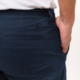 Close-up back view of a man wearing navy blue chino shorts, with one hand in his side pocket. Focus is on the right side showing the welt back pocket and the tonal navy blue embroidered dog logo above it