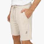 A close-up shot of a male model wearing beige athletic shorts. The shorts have a relaxed fit, an adjustable drawstring waist, and two large front pockets. The model's right hand is resting inside one of the pockets. A small embroidered logo of a sitting dog in black is visible near the hem on the left side. The fabric appears soft and textured. He pairs the shorts with a tucked-in white T-shirt, which is partially visible.