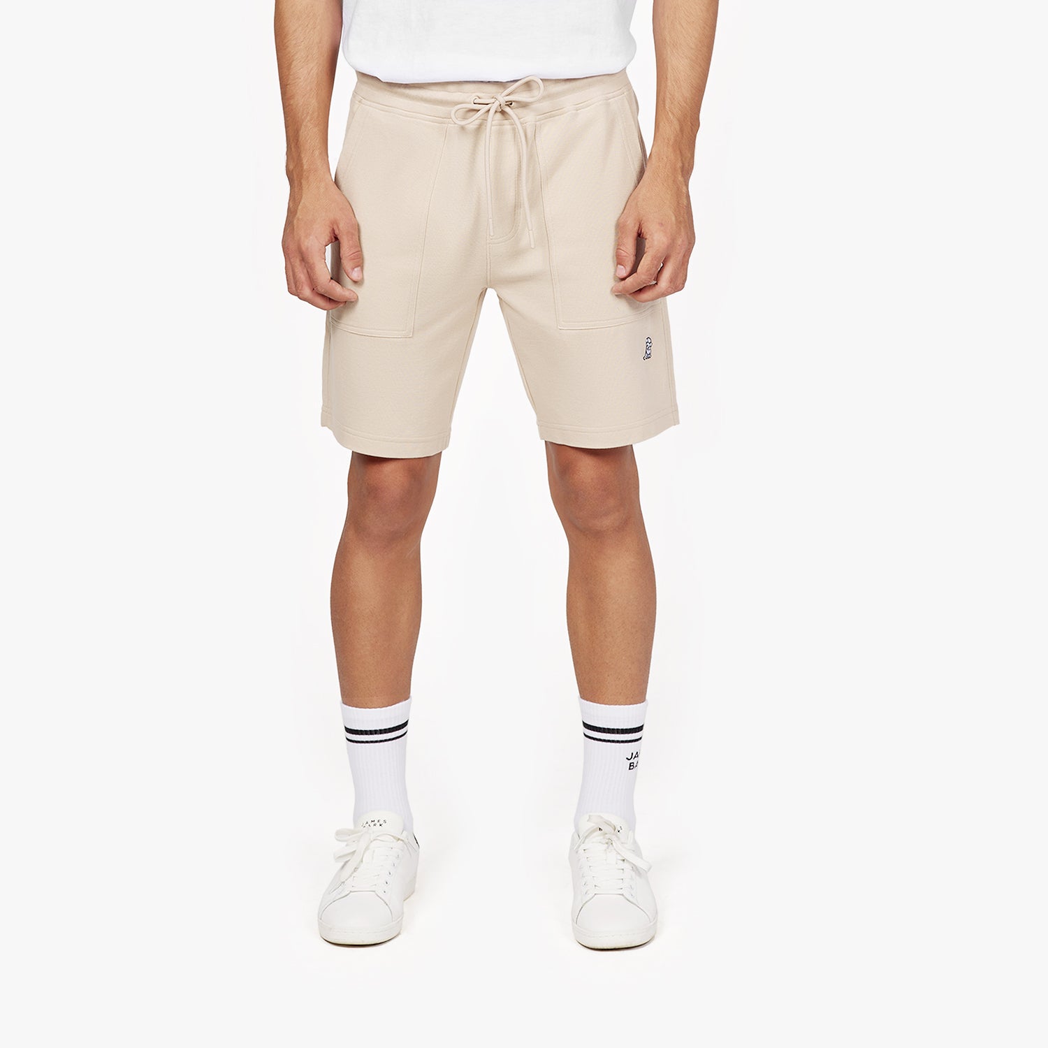 A male model is shown from the waist down, wearing beige athletic shorts against a plain white background. The shorts have a relaxed fit, an adjustable drawstring waist, and two large front pockets. A small embroidered logo of a sitting dog is visible near the hem on the left side. He pairs the shorts with white sneakers and white crew socks featuring black stripes and the brand name "JAMES BARK" near the top. His posture is neutral, with his hands resting at his sides.