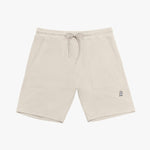 A pair of beige drawstring shorts laid flat against a white background. The shorts feature an elastic waistband with matching beige drawstrings tied in a bow. There are two large rectangular front pockets, stitched for a structured look. A small embroidered logo of a dog wearing a hat is positioned on the lower right leg. The shorts are made from a textured fabric, likely cotton or fleece, and have neatly finished hems