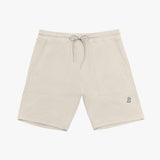 A pair of beige drawstring shorts laid flat against a white background. The shorts feature an elastic waistband with matching beige drawstrings tied in a bow. There are two large rectangular front pockets, stitched for a structured look. A small embroidered logo of a dog wearing a hat is positioned on the lower right leg. The shorts are made from a textured fabric, likely cotton or fleece, and have neatly finished hems