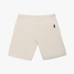 A pair of beige shorts laid flat against a white background, showcasing the back view. The shorts feature a clean, minimalist design with a single rectangular patch pocket on the right side, accented by a black woven brand label sewn into the seam. The waistband is elastic, and the fabric appears textured, likely cotton or fleece. The hem is neatly stitched, and the overall fit suggests a relaxed, comfortable style.