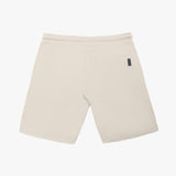 A pair of beige shorts laid flat against a white background, showcasing the back view. The shorts feature a clean, minimalist design with a single rectangular patch pocket on the right side, accented by a black woven brand label sewn into the seam. The waistband is elastic, and the fabric appears textured, likely cotton or fleece. The hem is neatly stitched, and the overall fit suggests a relaxed, comfortable style.