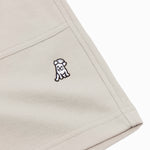 A close-up shot of a beige fabric, showcasing the texture and stitching details of a garment. The image highlights a small embroidered logo of a playful dog in black and white, positioned near the seam of a pocket. The fabric has a subtle woven texture, and the stitching along the edges appears precise and reinforced. The pocket design features a clean, structured appearance, contributing to the minimalist aesthetic of the garment