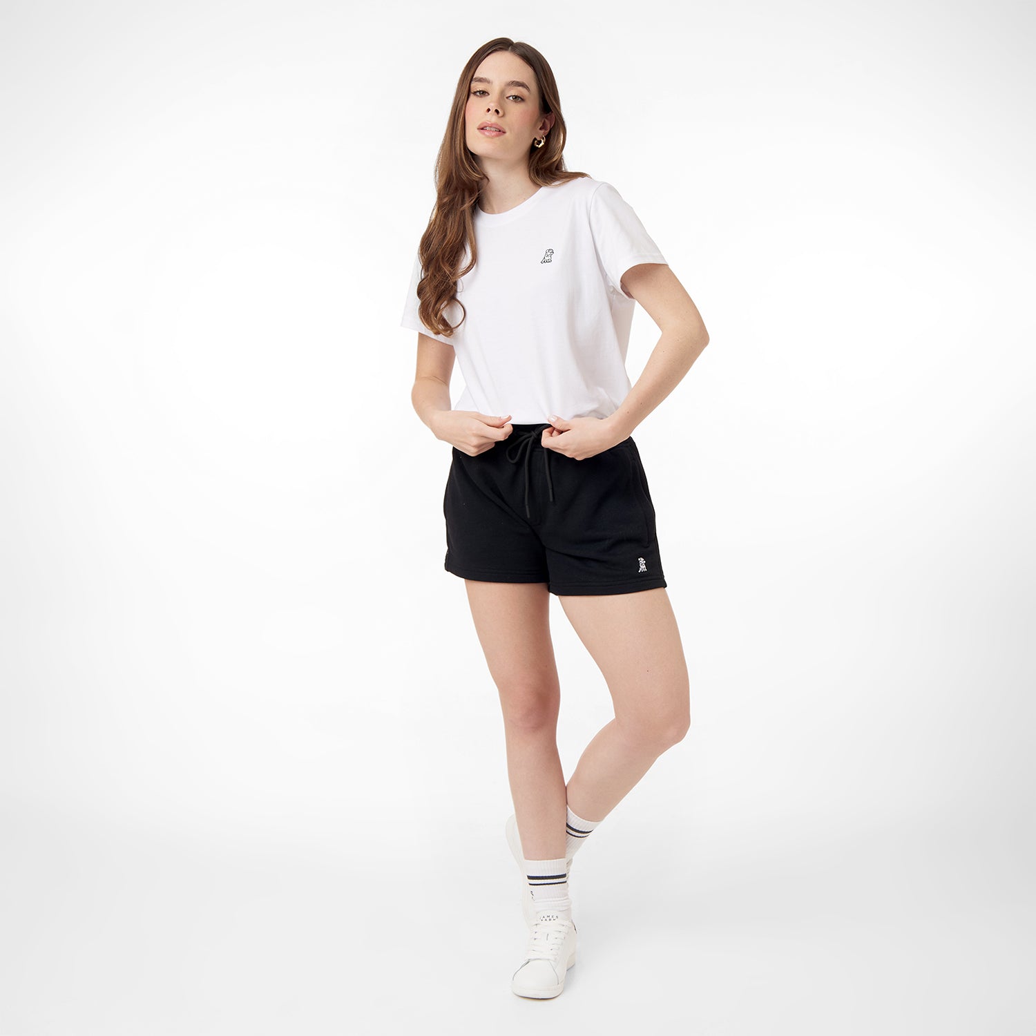 This image shows the model wearing the black shorts with the same logo on the lower left side, paired with a simple white t-shirt. She is standing casually, with both hands holding the waistband of the shorts. Her look is completed with white sneakers and striped socks, maintaining a relaxed, everyday style