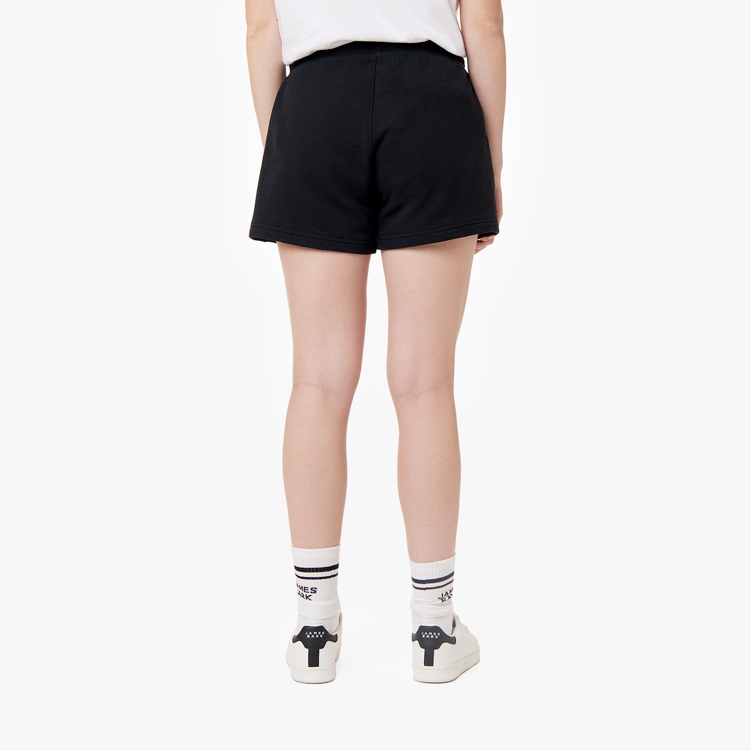 This image shows the same black shorts from the back. The model is still wearing the white t-shirt, white sneakers, and striped socks. The logo on the lower left side of the shorts is visible, maintaining the simple and relaxed style