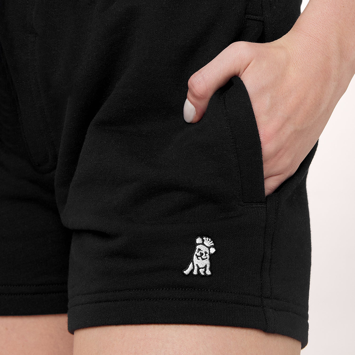 This close-up image shows the black shorts with the logo on the lower left side, similar to the previous ones. The logo is a small character with a raised hand and a hat. The model's hand is in the pocket, highlighting the relaxed fit of the shorts.