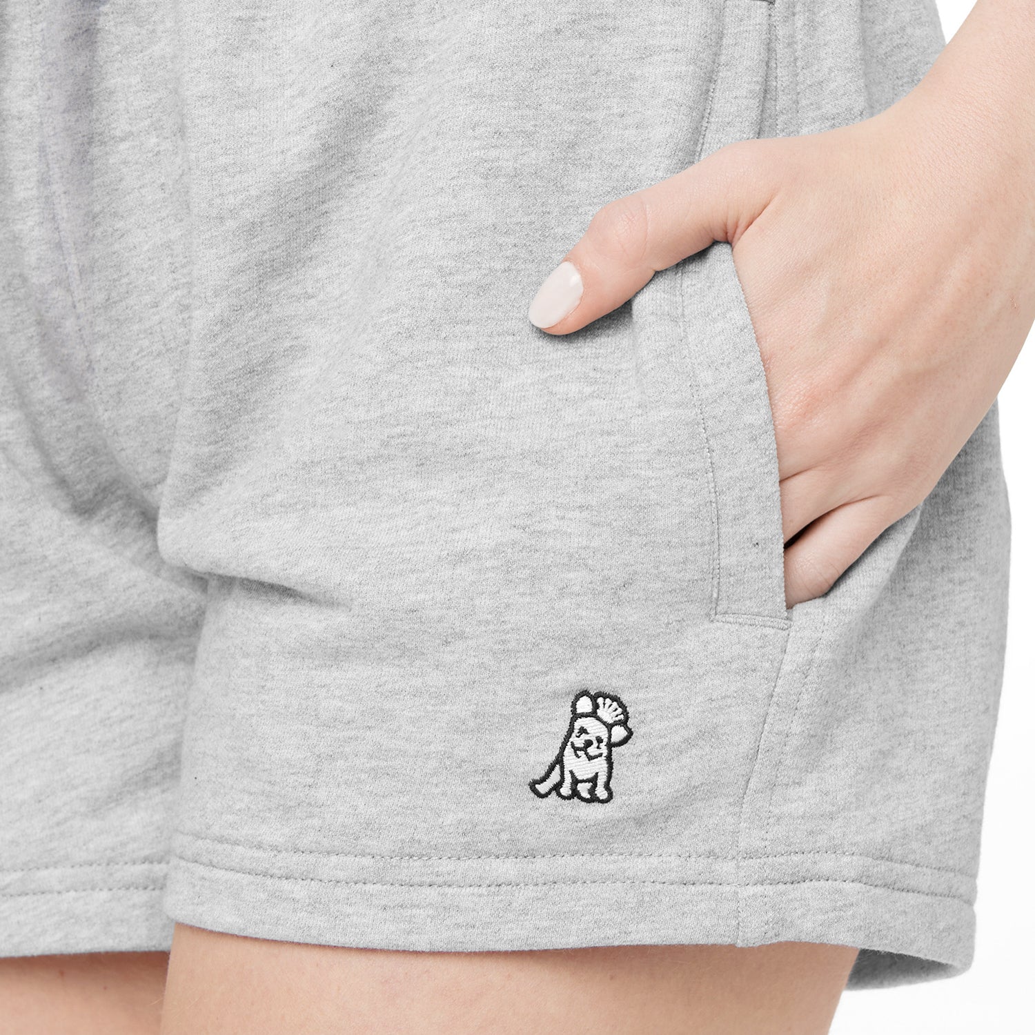 This image focuses on the logo on the gray shorts. The logo appears to be a small character, possibly a bear or similar creature, wearing a hat and with a hand raised. It is positioned on the lower part of the shorts, near the pocket