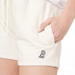 This image provides a close-up of the cream-colored shorts, showing the logo in greater detail. The logo features a character similar to the one on the gray shorts, still with a hat and hand raised. The model's hand is placed in the pocket, which emphasizes the relaxed style of the outfit.