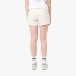 This image shows the same cream-colored shorts from the back, with the person still wearing the white t-shirt, white sneakers with blue accents, and striped socks. The logo on the shorts is visible on the left side, similar to the previous images