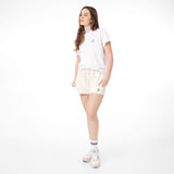 This image shows the same model wearing a white t-shirt with a small logo on the left chest area and the cream-colored shorts. The shorts have the same logo on the left side, and the model is wearing white sneakers with blue accents and striped socks. She's standing with one hand in her pocket, looking to the side. It gives off a casual, relaxed vibe.