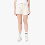 This image shows the same style of shorts, but in a different color—now in a light cream or off-white shade. The shorts still feature the same logo on the lower left side, and the model is wearing white sneakers and striped socks. The model's shirt is a simple white t-shirt, and she is standing in a neutral pose