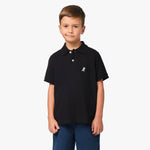 A young boy wearing a black polo shirt with a small embroidered logo on the chest, standing with his hands at his sides.