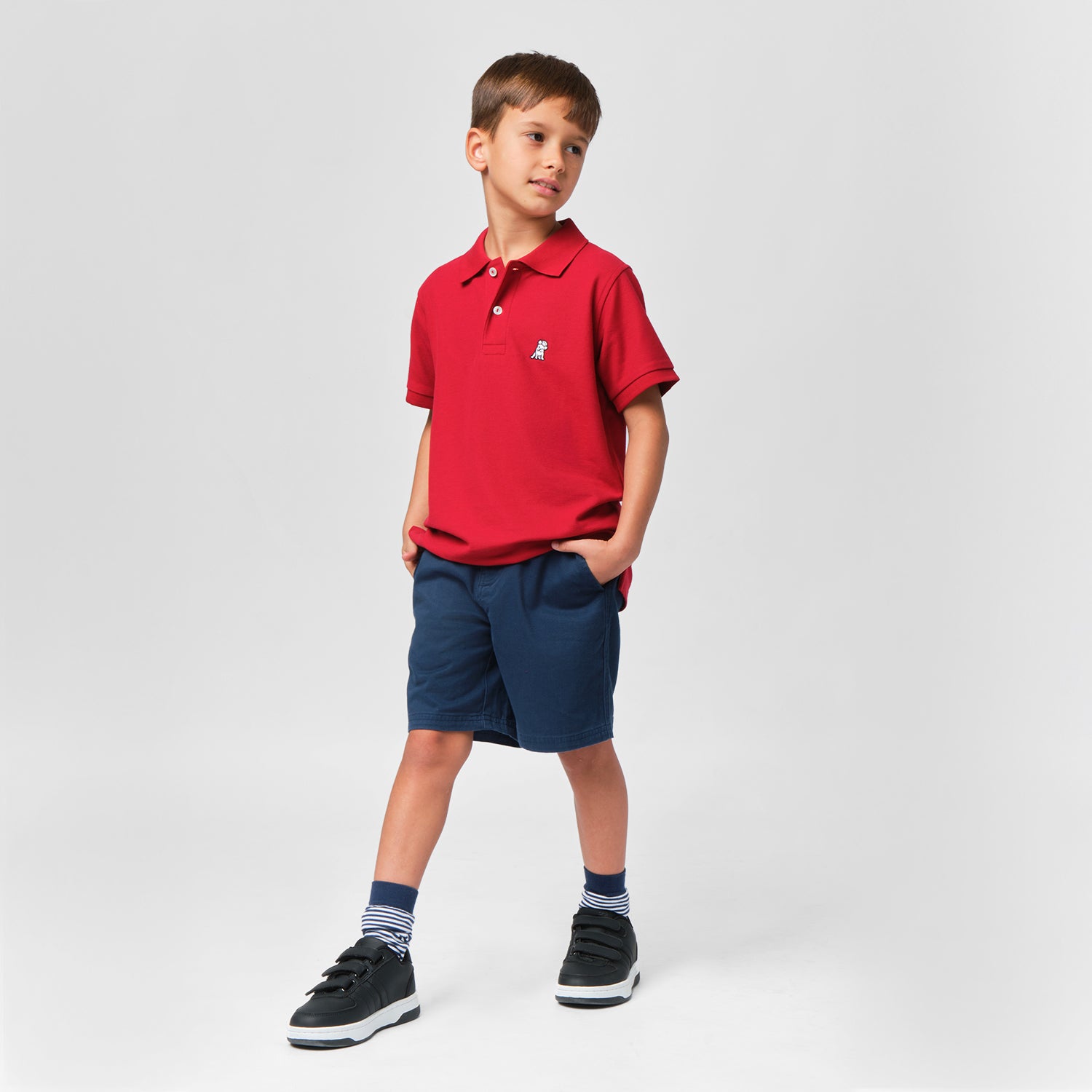 Full-body shot of the boy wearing the red polo shirt paired with navy blue shorts, standing and smiling with one hand in his pocket.