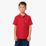 A young boy wearing a red polo shirt with a small blue embroidered logo on the chest, standing with his hands at his sides.