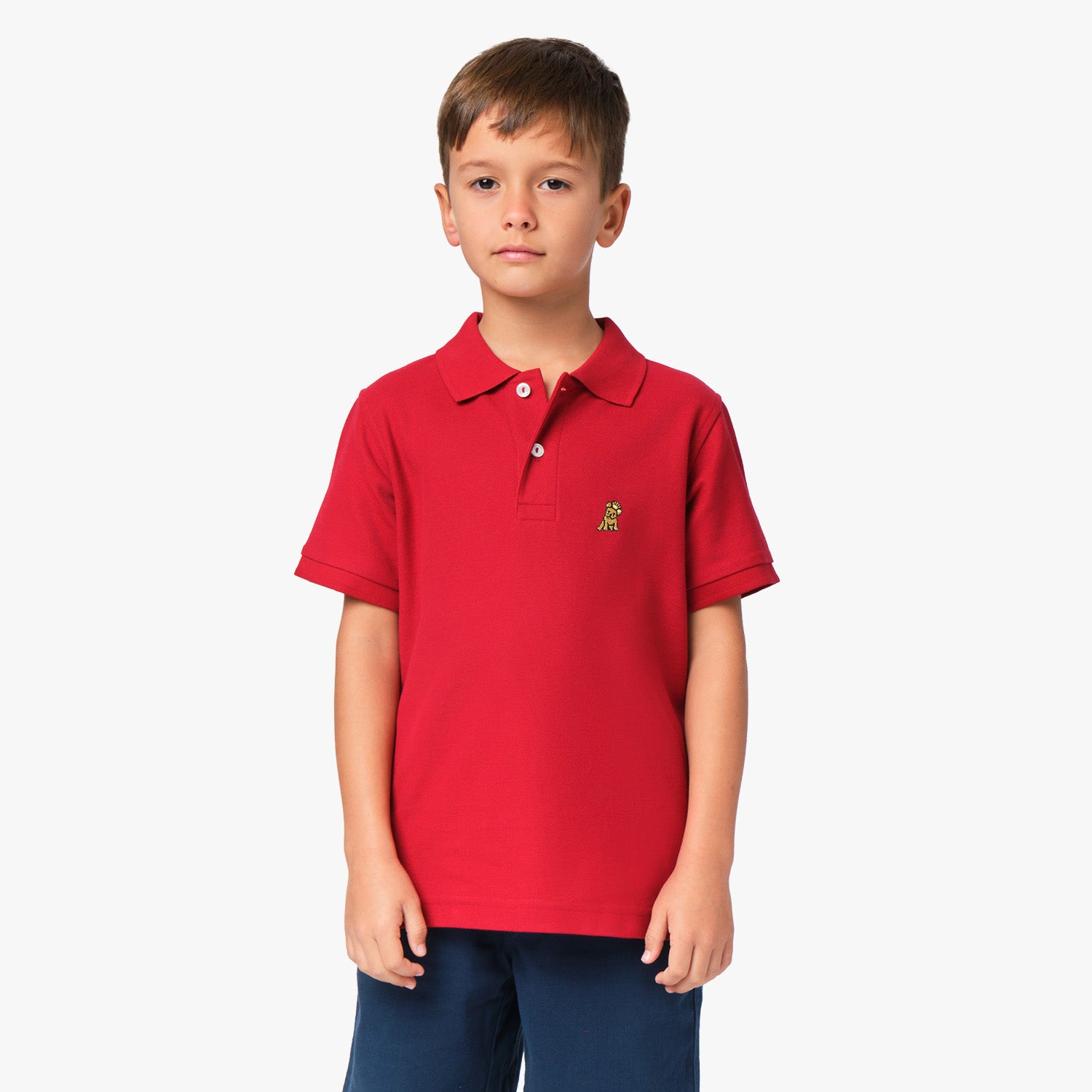 A young boy wearing a red polo shirt with a small gold embroidered logo on the chest, standing with his hands at his sides.