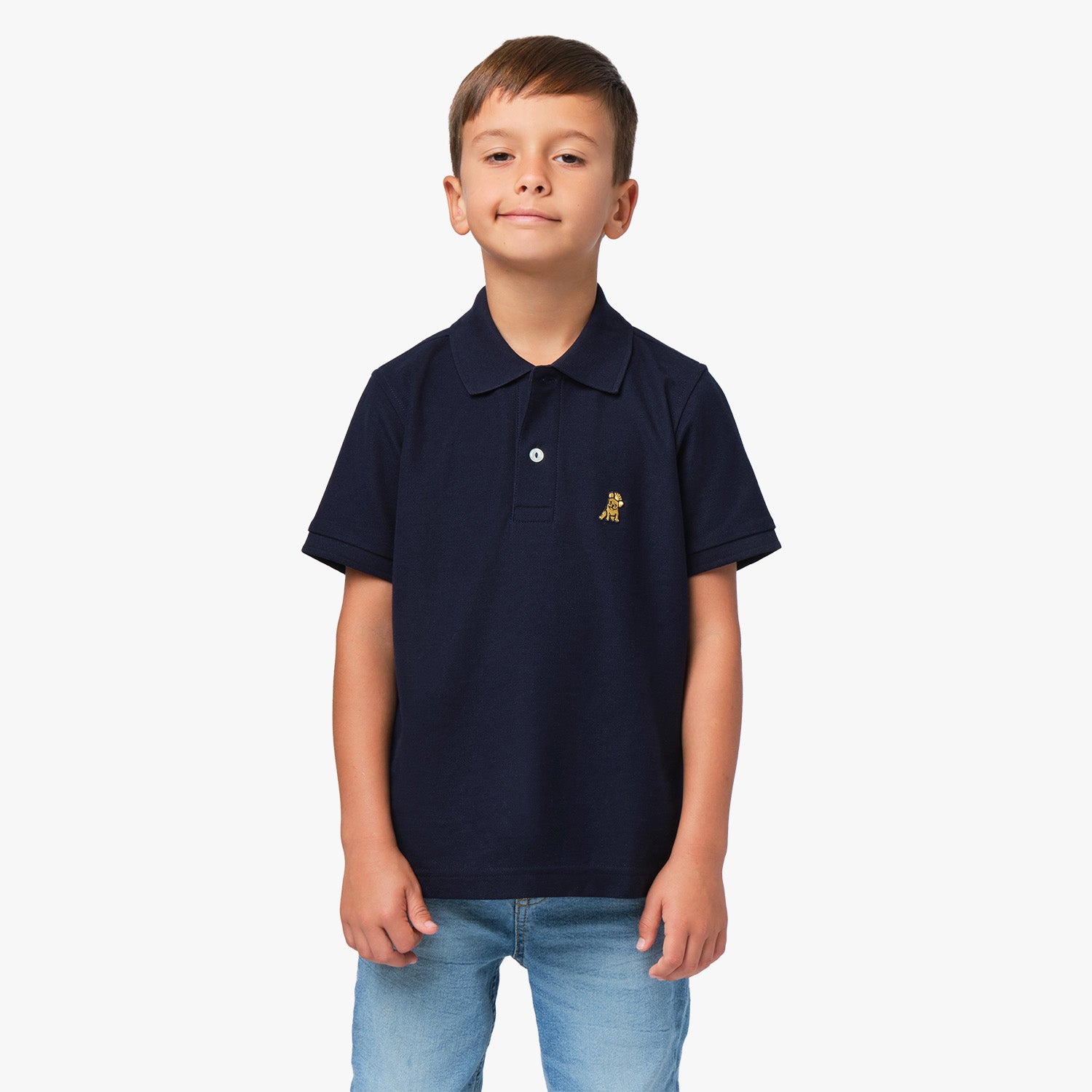 A young boy wearing a navy blue polo shirt with a small gold embroidered logo on the chest, standing and smiling with his hands at his sides.