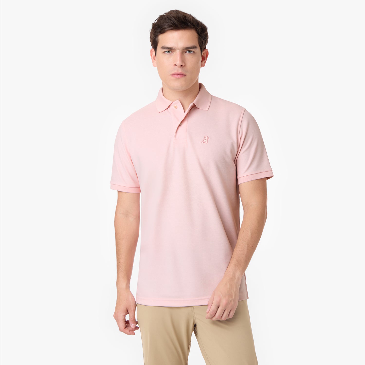 Male model wearing a pink polo shirt with a light pink embroidered dog logo and light grey pants, standing front view.