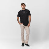 Male model wearing a black short-sleeve polo shirt with contrasting gray details on the collar, button placket, and sleeve cuffs. The polo is paired with beige slim-fit trousers and black sneakers with white soles. The model poses in a relaxed stance against a plain white background.