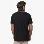 Rear view of a male model wearing a black short-sleeve polo shirt with contrasting gray details on the collar and sleeve cuffs. The polo has a smooth, structured fit. Paired with beige trousers for a modern and refined look