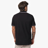 Rear view of a male model wearing a black short-sleeve polo shirt with contrasting gray details on the collar and sleeve cuffs. The polo has a smooth, structured fit. Paired with beige trousers for a modern and refined look