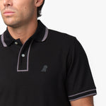 Close-up of a male model wearing a black short-sleeve polo shirt with contrasting gray details on the collar, button placket, and sleeve cuffs. The polo features a two-button design and a subtle embroidered dog logo on the chest, blending seamlessly with the fabric