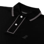 A close-up of a black polo shirt featuring gray trim along the collar and placket. The placket has two white buttons, and the brand label "James Bark Est. 2015" is visible inside the collar. The shirt’s textured fabric is accompanied by a subtle embossed dog logo on the left chest, blending into the black fabric.