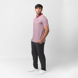 A male model poses in a side profile wearing a slim-fit, short-sleeve polo shirt with a fine red and white stripe pattern. The collar is burgundy with a light pink trim, and the sleeve cuffs have matching accents. He pairs the polo with dark gray joggers and white sneakers. His stance is relaxed, with his hands in his pockets, and he gazes slightly away from the camera against a plain light gray background