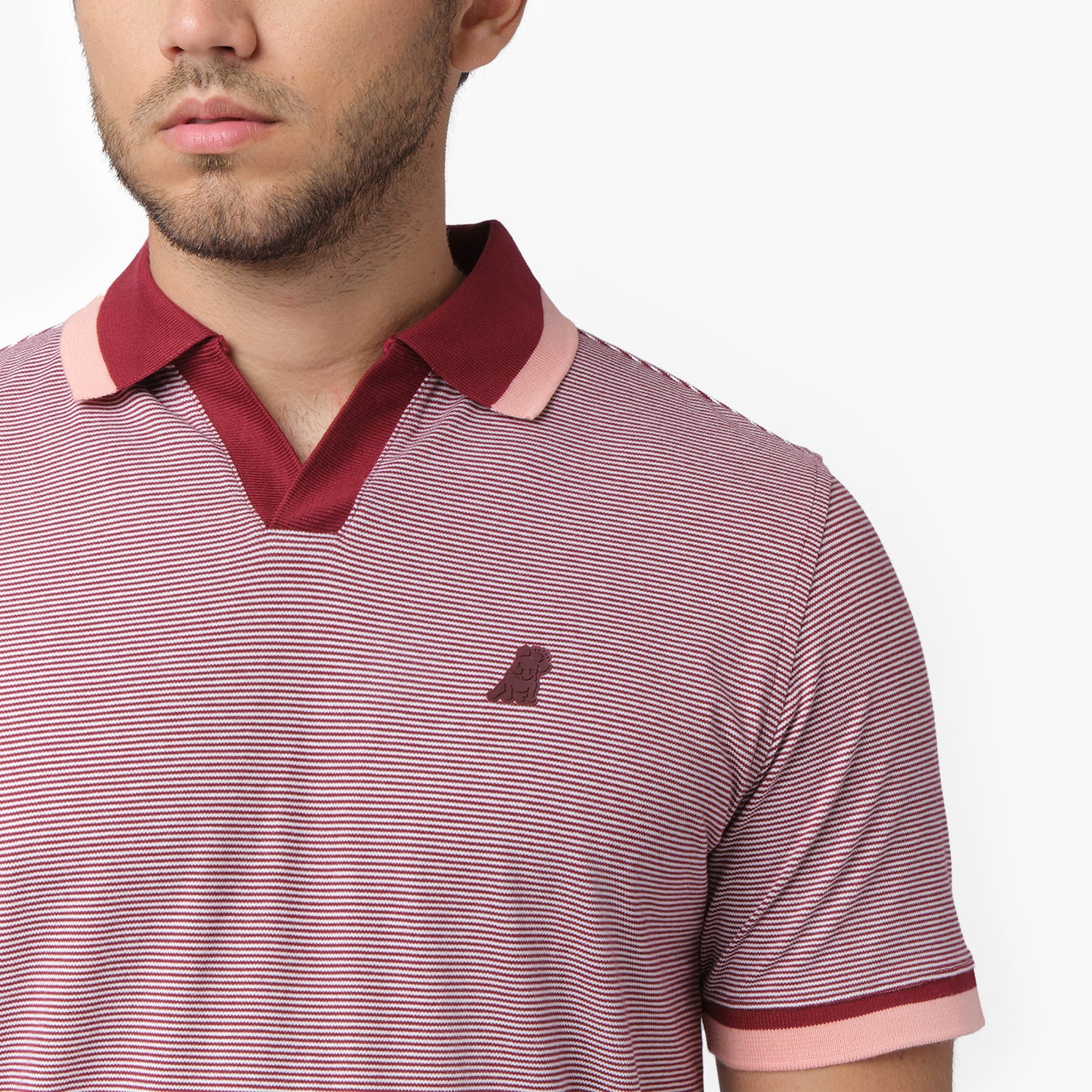 A close-up of a male model wearing a short-sleeve polo shirt with a fine red and white stripe pattern. The collar has a contrasting burgundy and light pink trim, matching the sleeve cuffs. A small embroidered logo of a dog is visible on the left chest area. The fabric appears textured, and the model’s face is partially visible, showing his beard and neutral expression