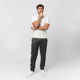 Male model wearing a white short-sleeve polo shirt paired with dark gray fitted trousers and white sneakers. The polo features a V-neck collar and ribbed sleeve cuffs. The model poses against a plain white background with a thoughtful stance, hand resting on his chin.