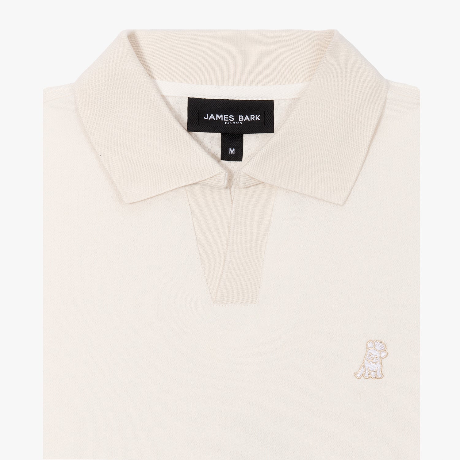 A close-up of a cream-colored polo shirt from the brand "JAMES BARK." The ribbed collar and V-neck opening give it a refined look. A black label inside the collar displays the brand name, establishment year "Est. 2015," and size "M." The shirt’s fabric appears soft and textured. On the left chest, there's a small embroidered dog logo in matching cream thread, subtly blending with the overall design