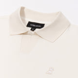 A close-up of a cream-colored polo shirt, highlighting the ribbed collar and short V-neck opening. The black neck label displays the brand name "JAMES BARK" with "Est. 2015" beneath it, along with the size "M." The fabric has a smooth texture. A small embroidered logo of a dog in a subtle cream-colored thread is positioned on the left chest area, blending elegantly with the fabric. The material appears soft and breathable