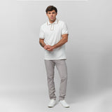 Male model wearing a white short-sleeve polo shirt with beige contrast details on the collar, button placket, and sleeve cuffs. The polo is paired with slim-fit light gray trousers and white sneakers. The model poses against a plain gray background with a relaxed stance