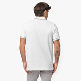 Rear view of a male model wearing a white short-sleeve polo shirt with beige contrast details on the collar and sleeve cuffs. The polo has a smooth, structured fit. Paired with light gray trousers for a clean, modern look