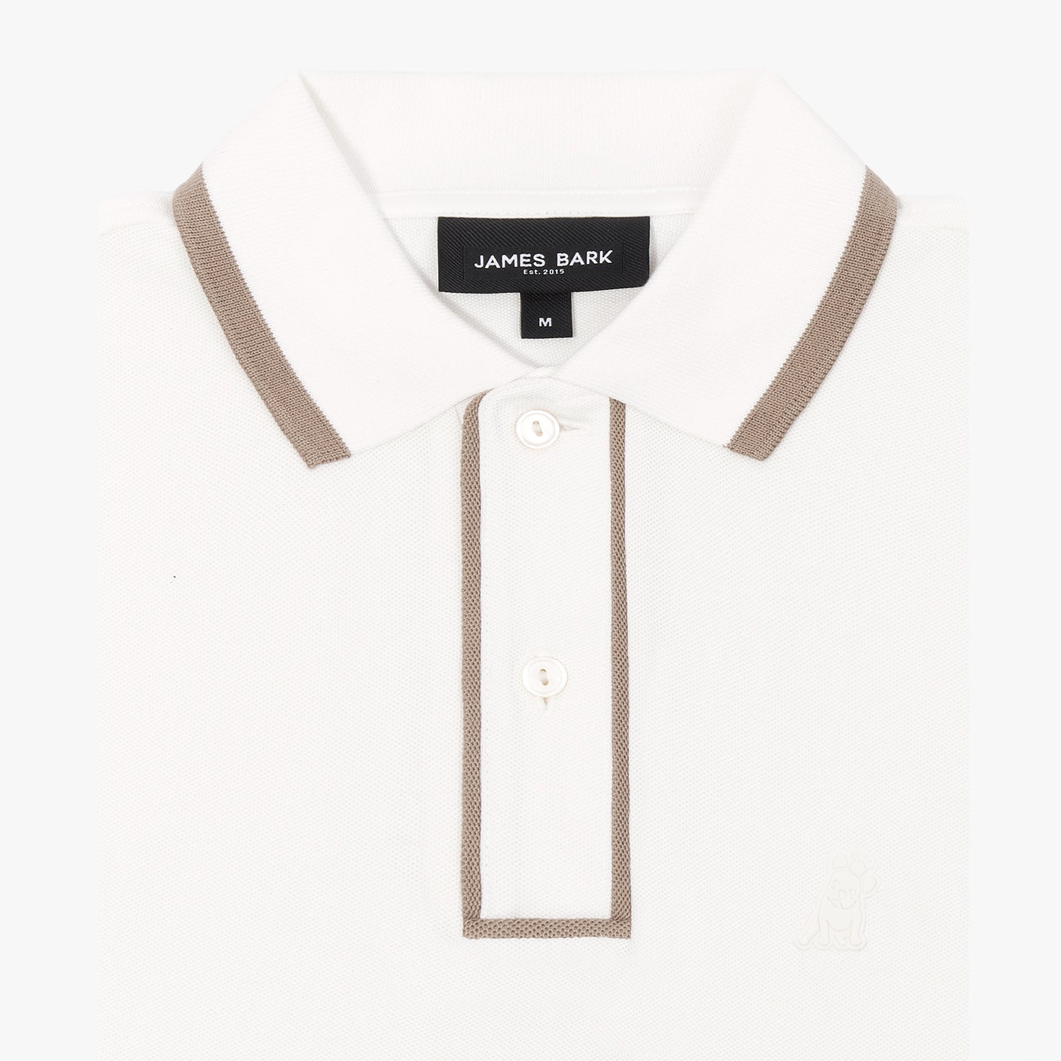 A close-up of a white polo shirt with brown trim on the collar and placket. The fabric has a subtle textured weave. The two-button closure is visible, along with a black label on the inside of the collar displaying the brand name "James Bark" and the establishment year "Est. 2015." A faint embroidered logo of a dog is subtly placed on the lower right side of the shirt