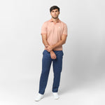 Male model wearing a peach-colored short-sleeve polo shirt paired with blue drawstring trousers and white sneakers. The polo features a V-neck collar, ribbed sleeve cuffs, and a small embroidered white dog logo on the chest. The model poses against a plain white background with a relaxed stance