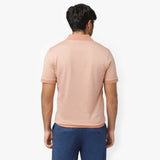 Rear view of a male model wearing a peach-colored short-sleeve polo shirt. The polo features a classic collar, ribbed sleeve cuffs, and a smooth, fitted design. Paired with blue trousers for a stylish contrast