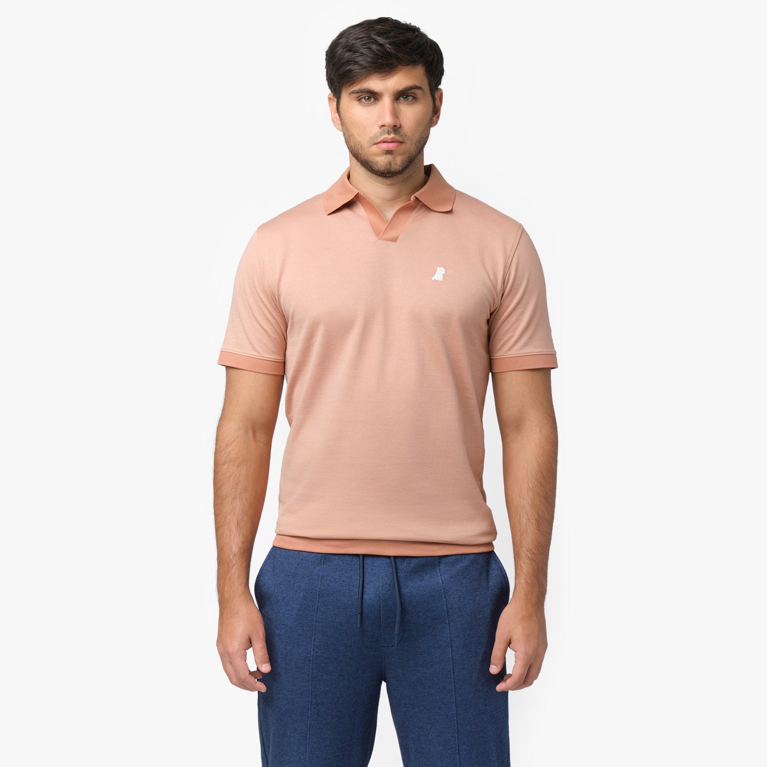 Male model wearing a peach-colored short-sleeve polo shirt with a V-neck collar, ribbed sleeve cuffs, and a small embroidered white dog logo on the chest. Paired with blue drawstring trousers for a stylish and casual look