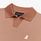 A close-up of a brown polo shirt, showcasing the ribbed collar and short V-neck opening. The black neck label displays the brand name "JAMES BARK" with "CLOSET" beneath it, along with the size "M." The fabric has a finely textured weave. A small embroidered logo of a dog in white thread is positioned on the left chest area, standing out against the brown fabric. The material appears soft and breathable