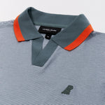 Close-up of a striped polo shirt with a micro-stripe pattern in blue and white. The ribbed collar is deep green, accented with a burnt orange stripe along the edge. The open placket gives the shirt a modern, relaxed feel. The James Bark brand label and size "M" are visible inside the collar. On the left chest, a tonal rubberized dog logo adds a subtle yet distinct branding detail.