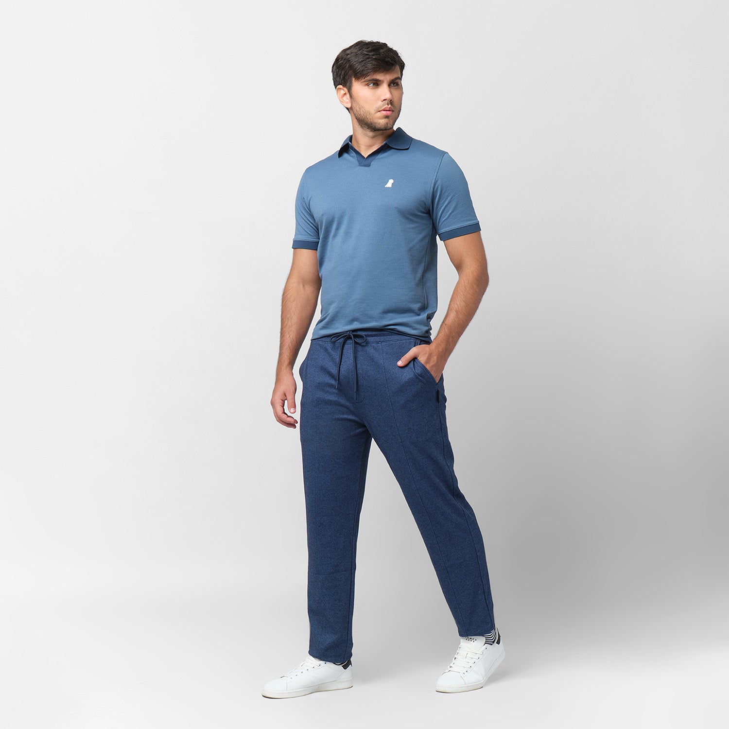 Male model wearing a blue short-sleeve polo shirt paired with relaxed-fit navy blue drawstring trousers and white sneakers. The polo features a V-neck collar, ribbed sleeve cuffs, and a small embroidered white dog logo on the chest. The model poses against a plain white background