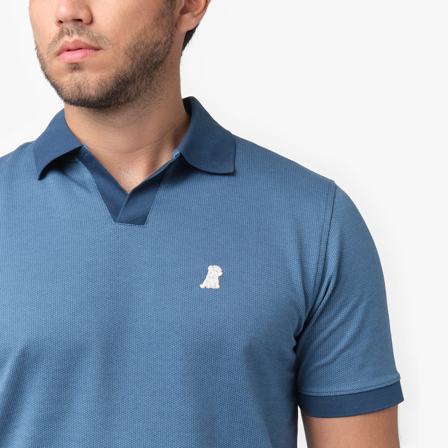 Close-up of a male model wearing a blue short-sleeve polo shirt. The polo features a textured fabric, a V-neck collar with a contrasting inner lining, ribbed sleeve cuffs, and a small embroidered white dog logo on the chest