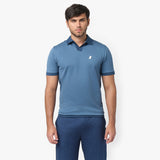 Male model wearing a blue short-sleeve polo shirt with a relaxed fit. The polo features a subtle V-neck collar, ribbed sleeve cuffs, and a small embroidered white dog logo on the chest. Paired with matching blue trousers