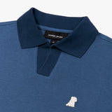 Close-up of a navy blue polo shirt from "JAMES BARK," showcasing its textured fabric and contrasting darker blue ribbed collar. The V-neck opening features a reinforced darker blue placket. A small embroidered white dog logo is positioned on the left chest. The inside collar label displays the brand name and the shirt size "M" on a black background.