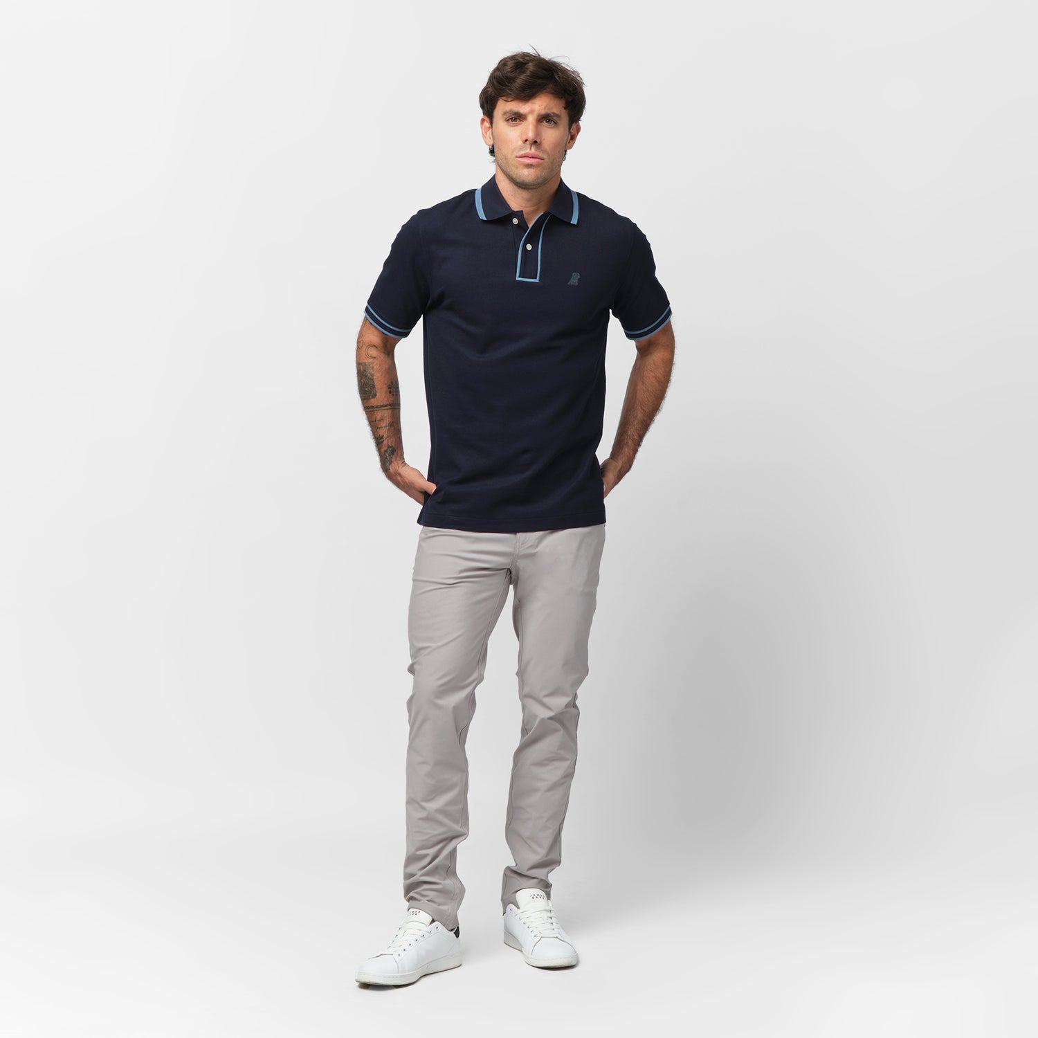 Full-body shot of a male model wearing a navy blue polo shirt with light blue accents on the collar and sleeve cuffs, paired with slim-fit light gray pants and white sneakers. He stands against a plain white background with a relaxed posture, slightly resting his hands on his hips