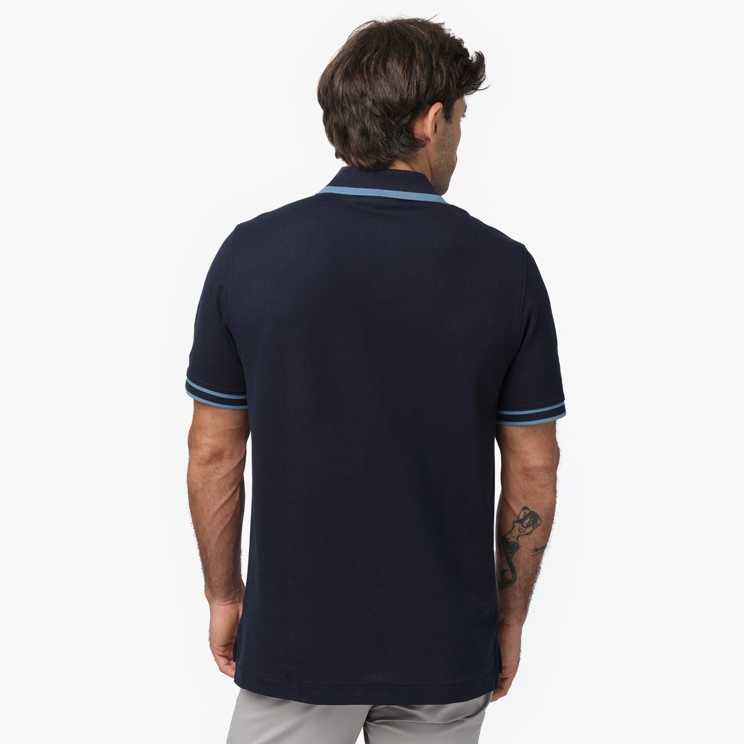 Rear view of a male model wearing a navy blue short-sleeve polo shirt with contrasting light blue details on the collar and sleeve cuffs. The polo has a structured fit and is paired with light gray trousers for a sleek and casual look